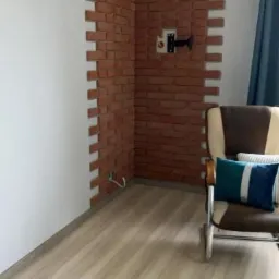 Fragment ściany wyłożony dekoracyjną cegłą w rogu pokoju, z widocznym uchwytem na telewizor, jasnymi panelami podłogowymi i fotelem z poduszką w tle.