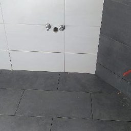 Rem-Bud - Świeżo ułożone, ciemnoszare płytki podłogowe w łazience, kontrastujące z białymi płytkami ściennymi. Widoczne otwory na armaturę. Częściowo ułożone płytki na ścianie bocznej.