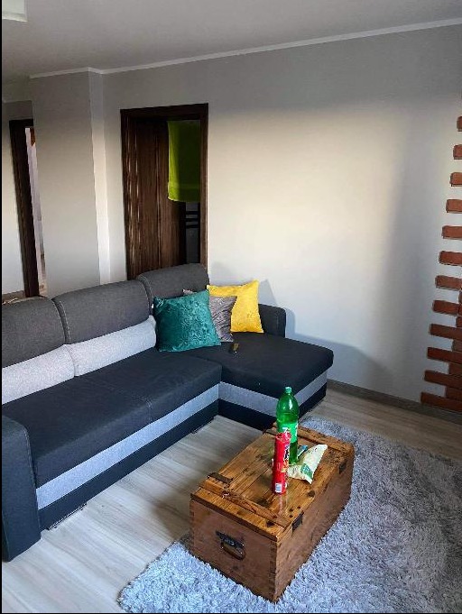 Jasnoszary narożnik z zieloną i żółtą poduszką, drewniana skrzynia pełniąca funkcję stolika kawowego z butelką napoju i przekąskami, ściana w odcieniach szarości z fragmentem cegieł dekoracyjnych.