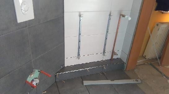 W trakcie montażu szarej, wielkoformatowej glazury podłogowej w łazience, widoczna poziomica, metalowe profile mocujące na ścianie, instalacja wodna i fragment grzejnika.