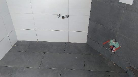 Świeżo ułożone, ciemnoszare płytki podłogowe w łazience, kontrastujące z białymi płytkami ściennymi. Widoczne otwory na armaturę. Częściowo ułożone płytki na ścianie bocznej.