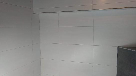 Ściana wyłożona jasnymi, prostokątnymi płytkami ceramicznymi z ciemnoszarą fugą, częściowo widoczny ciemny element wyposażenia w prawym dolnym rogu.