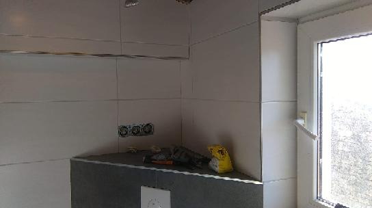 Ściana łazienki wyłożona jasnymi, poziomymi płytkami z metalowymi listwami, widoczne gniazdka elektryczne, fragment okna i narzędzia na szarej półce, w tym żółty worek z zaprawą.