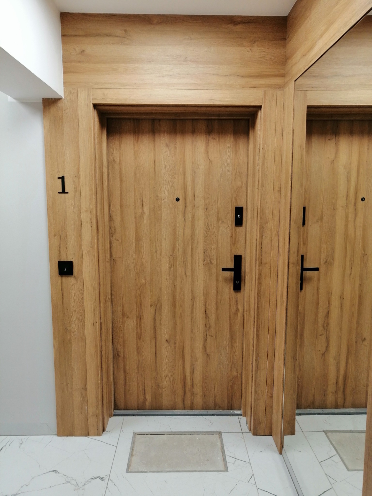 Nowoczesne drzwi wejściowe z numerem '1' w mieszkaniu we Wrocławiu, widoczne w lustrzanym odbiciu, z czarną klamką i wizjerem, dopasowane do drewnianej boazerii.