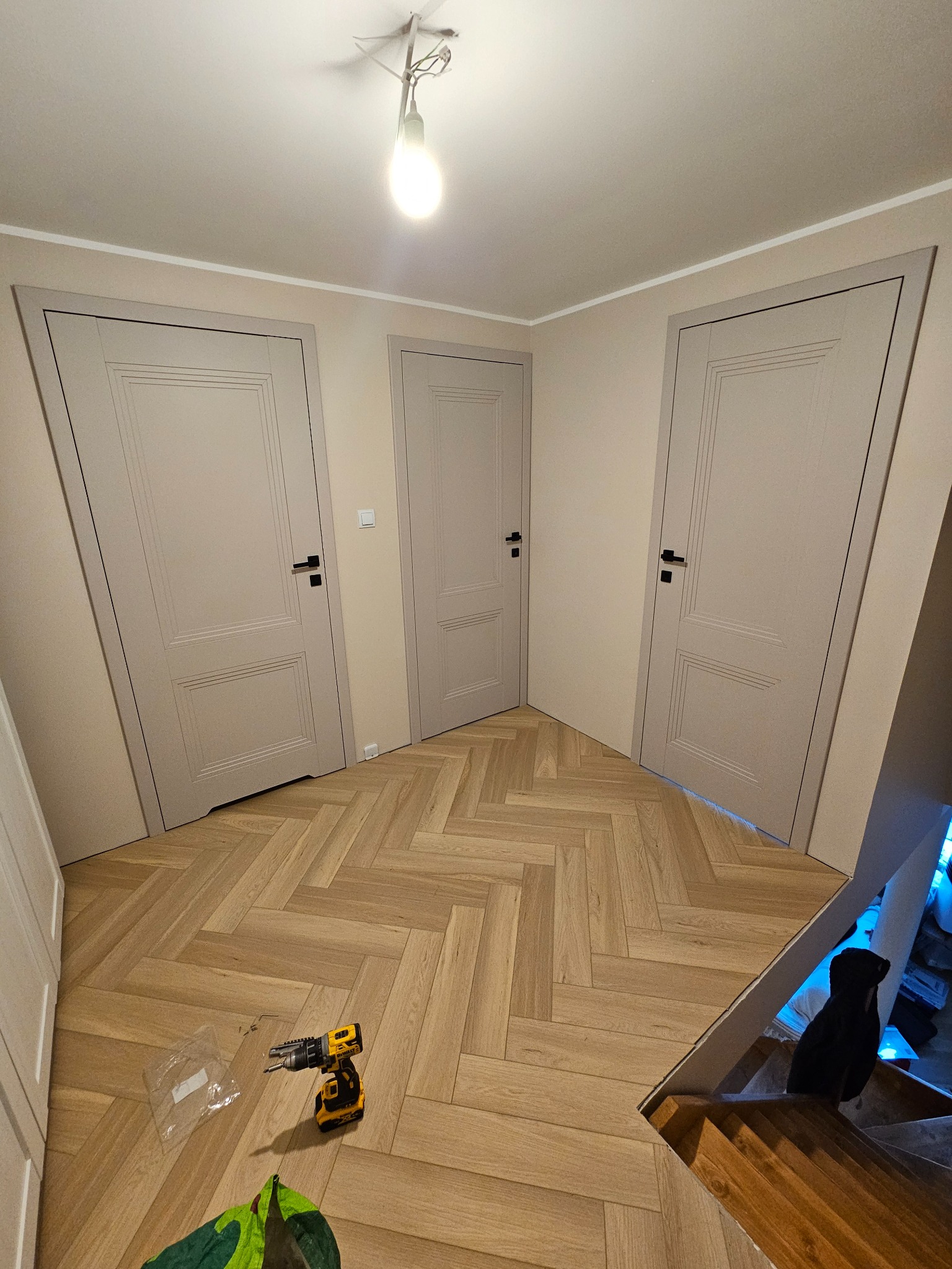 Trzy szare, panelowe drzwi wewnętrzne z czarnymi klamkami, zamontowane w nowym domu. Na podłodze parkiet w jodełkę i wkrętarka. Widok z dołu schodów.