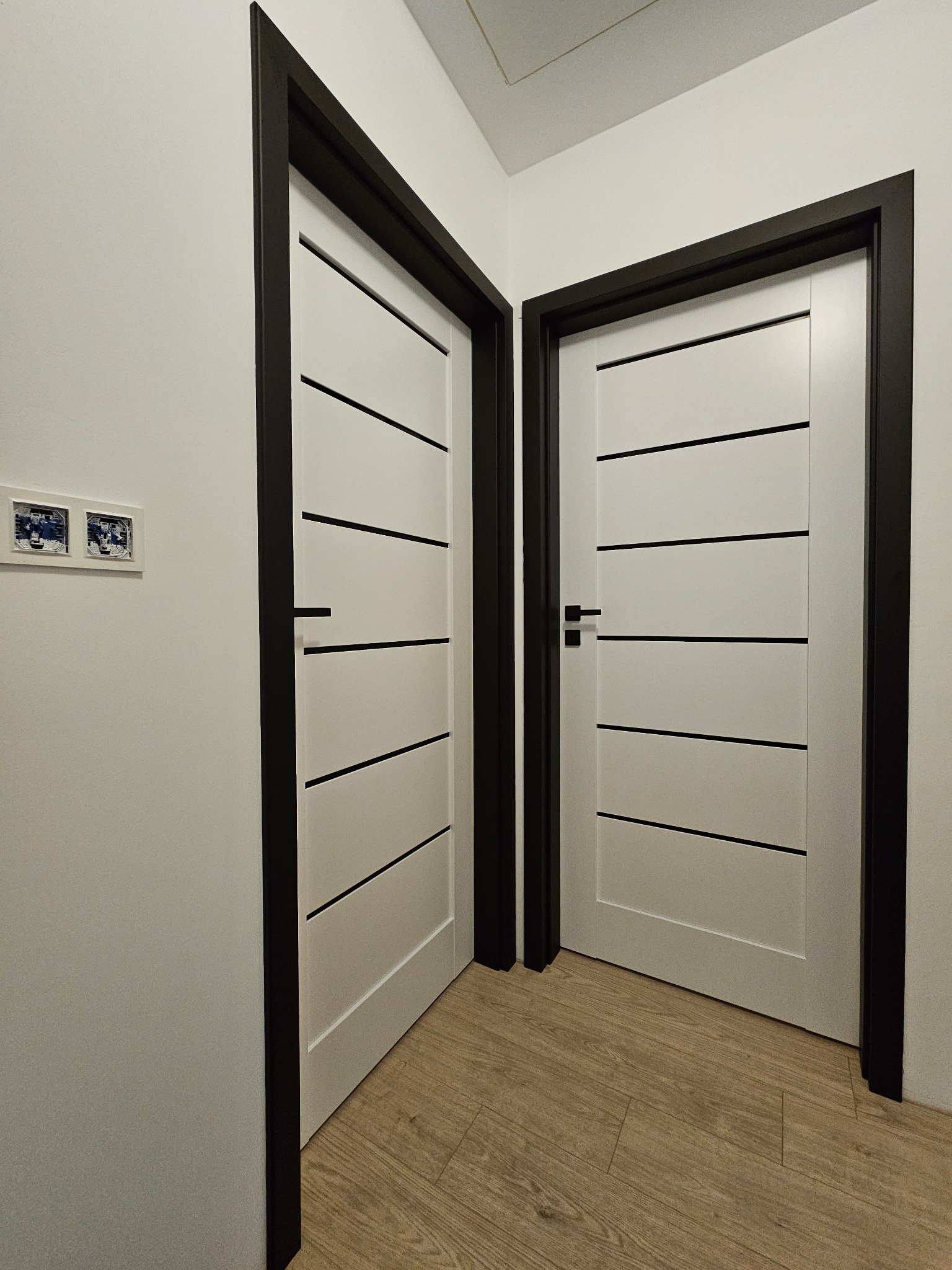 Dwa białe, nowoczesne drzwi z czarnymi ramami i poziomymi paskami, zamontowane w białym wnętrzu z drewnianą podłogą. Na ścianie widoczne zdjęcia w ramce.