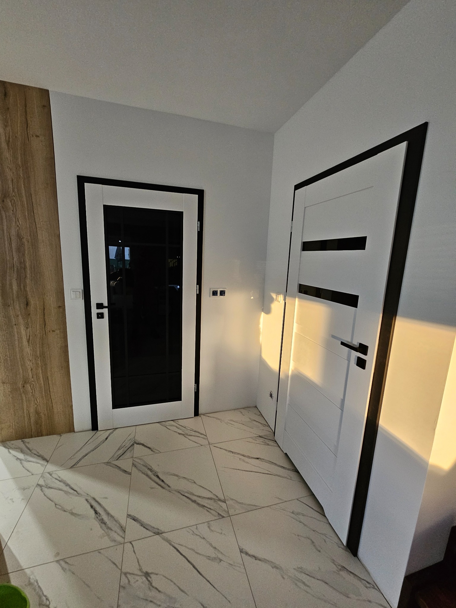Dwa białe skrzydła drzwiowe z czarnymi akcentami w nowoczesnym wnętrzu. Jedne drzwi z przeszkleniem, drugie z poziomymi wstawkami. Marmurowe płytki podłogowe.