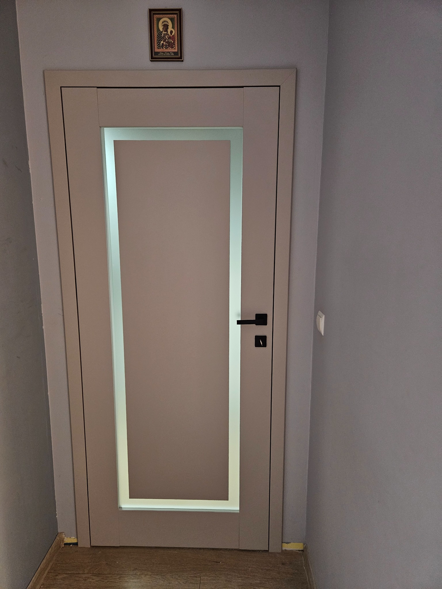 Zamknięte, beżowe drzwi wewnętrzne z prostokątnym przeszkleniem, czarną klamką i ikoną nad futryną, na tle szarej ściany i drewnianej podłogi.