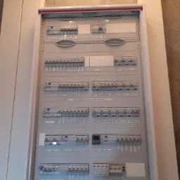 Rozdzielnica elektryczna w obudowie, widoczne bezpieczniki i wyłączniki z opisami, zamontowana na ścianie w surowym stanie.