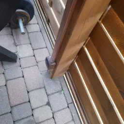 Brązowy fotokomórka zamontowana na słupku drewnianego ogrodzenia panelowego, widoczna kostka brukowa i fragment obciążenia do ćwiczeń.