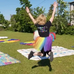 Animator w tęczowej spódniczce, w podskoku z uniesionymi rękami na trawie, z tyłu widoczna koszulka z napisem 'Animacje Pauli' i numerem telefonu, w tle drzewa i budynek.