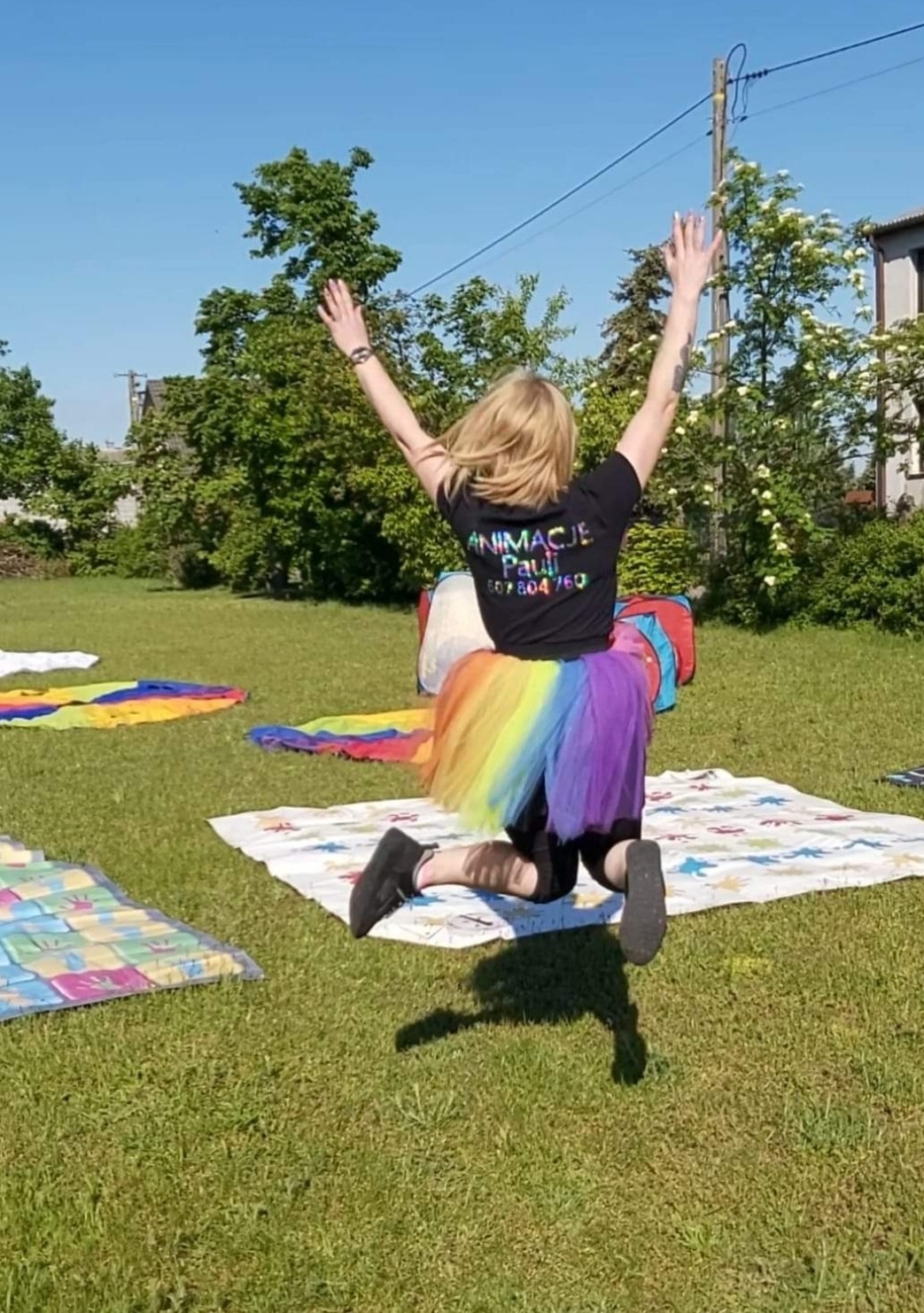 Animator w tęczowej spódniczce, w podskoku z uniesionymi rękami na trawie, z tyłu widoczna koszulka z napisem 'Animacje Pauli' i numerem telefonu, w tle drzewa i budynek.