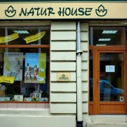 Fasada budynku z szyldem 'Natur House', okno wystawowe z plakatami i produktami, brązowe drzwi wejściowe, rynna odprowadzająca wodę.