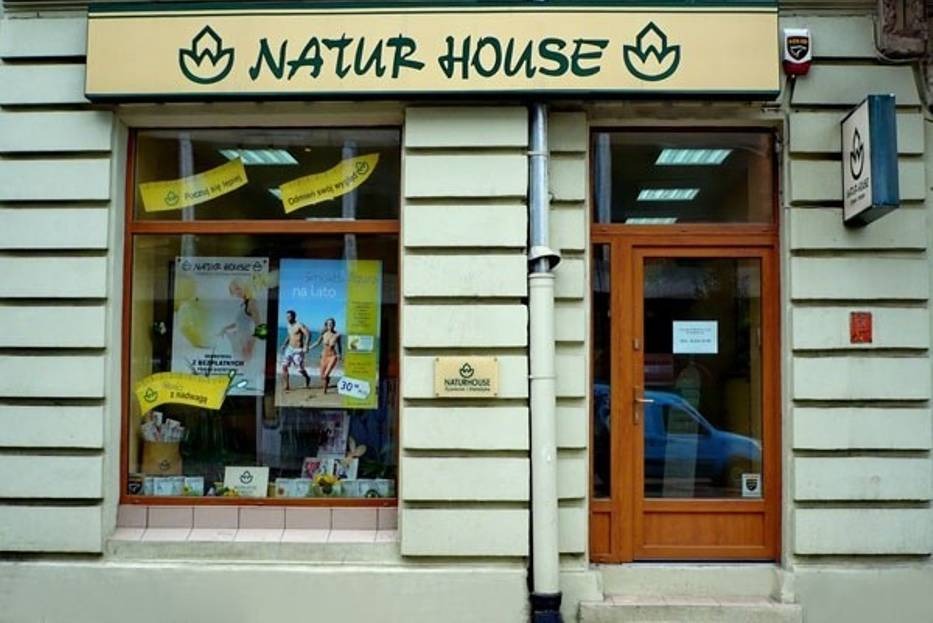 Fasada budynku z szyldem 'Natur House', okno wystawowe z plakatami i produktami, brązowe drzwi wejściowe, rynna odprowadzająca wodę.