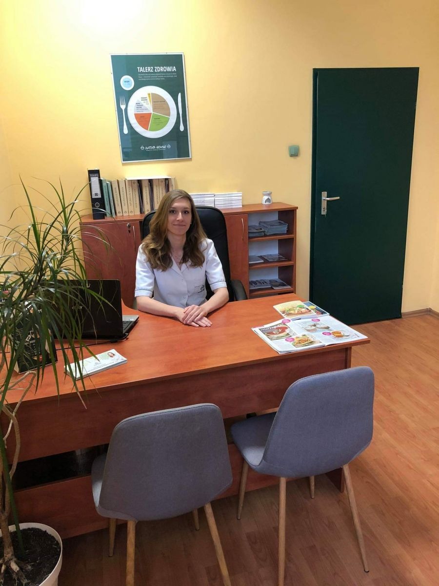 Kobieta w białym uniformie siedzi za biurkiem w gabinecie z plakatem 'Talerz Zdrowia' na ścianie, laptopem i ulotkami na blacie.
