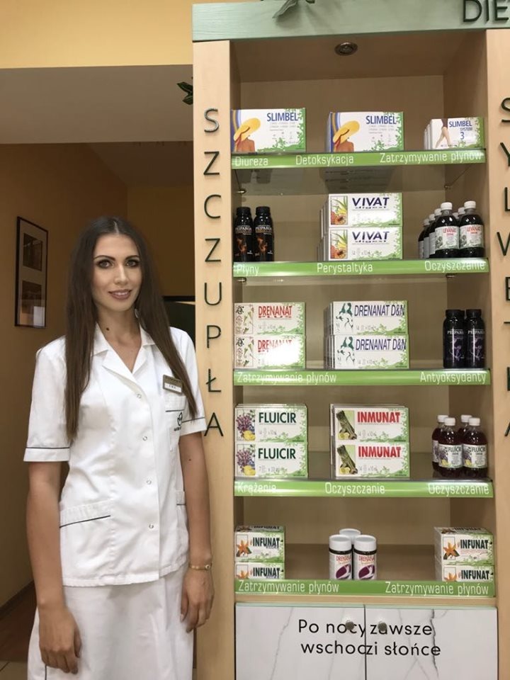 Kobieta w białym uniformie, prawdopodobnie pracownik gabinetu, stoi obok regału z suplementami diety i produktami ziołowymi, promując zdrowy tryb życia.