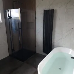 Nowoczesna łazienka z czarną kabiną prysznicową typu walk-in, grzejnikiem drabinkowym i białą wanną wypełnioną wodą, wykończona płytkami imitującymi marmur i betonową podłogą.