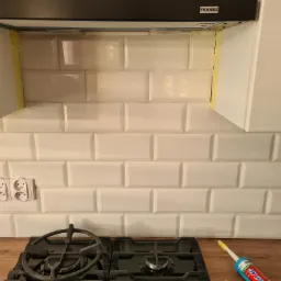 Białe, prostokątne płytki ułożone nad kuchenką gazową, częściowo zabezpieczone taśmą malarską. Widoczny okap marki Franke, gniazdka elektryczne oraz tuba z silikonem i akcesoria montażowe.