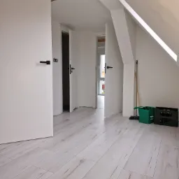 Poddasze po remoncie w Trzebiatowie: białe ściany, jasna podłoga imitująca drewno, otwarte białe drzwi z czarnymi klamkami, widoczne skrzynki i miotła w rogu.