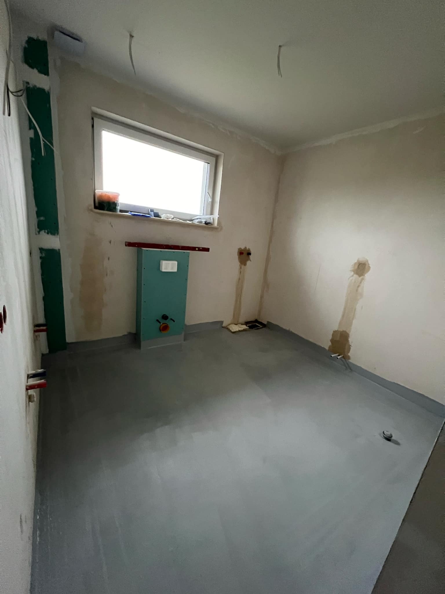 Surowy stan łazienki w trakcie prac: wylewka na podłodze, zabudowa stelaża WC z płyt karton-gips, wyprowadzone instalacje elektryczne i wodno-kanalizacyjne w ścianach, widoczne kable oświetleniowe...