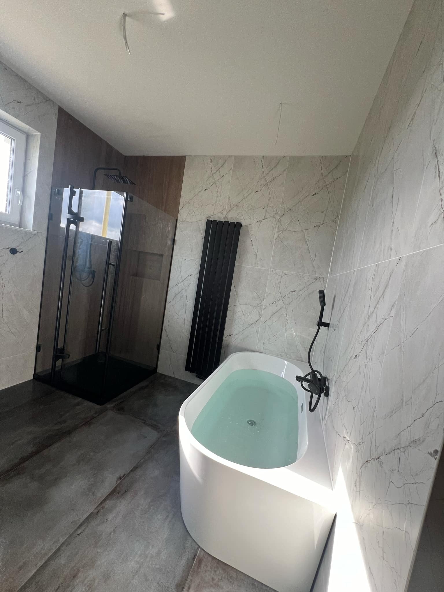 Nowoczesna łazienka w Trzebiatowie z białą wanną wypełnioną wodą, czarną kabiną prysznicową i grzejnikiem drabinkowym na tle marmurowych płytek ściennych i szarej podłogi.