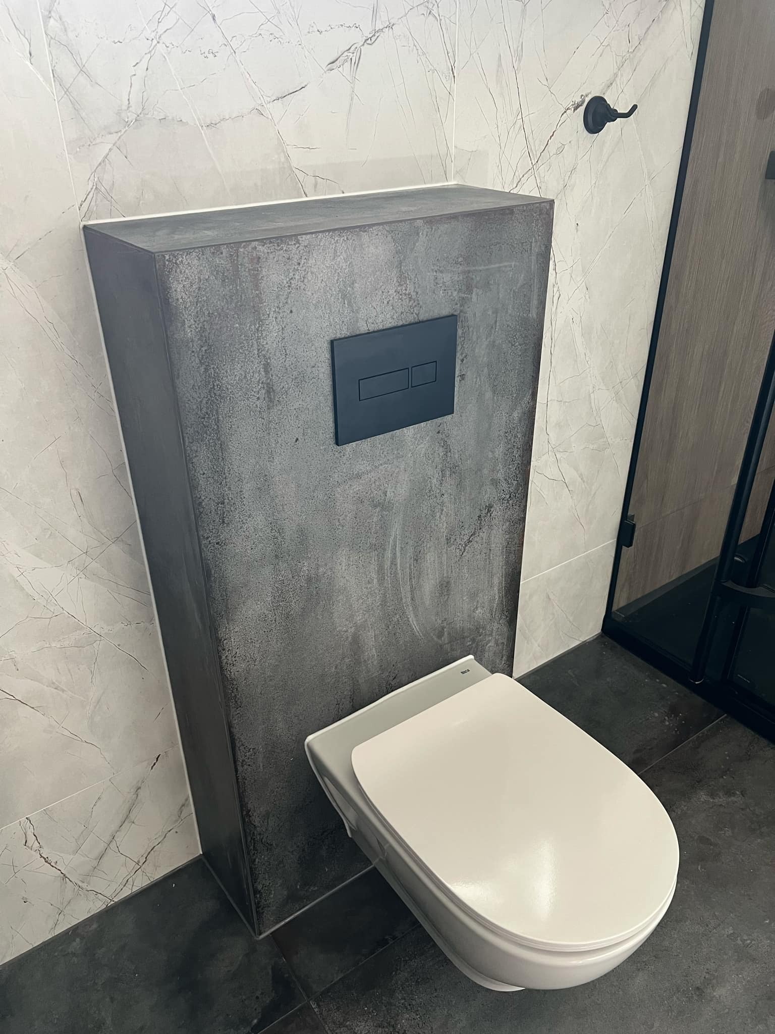 Podwieszana toaleta z szarą zabudową w łazience z marmurowymi płytkami na ścianach i czarną armaturą.