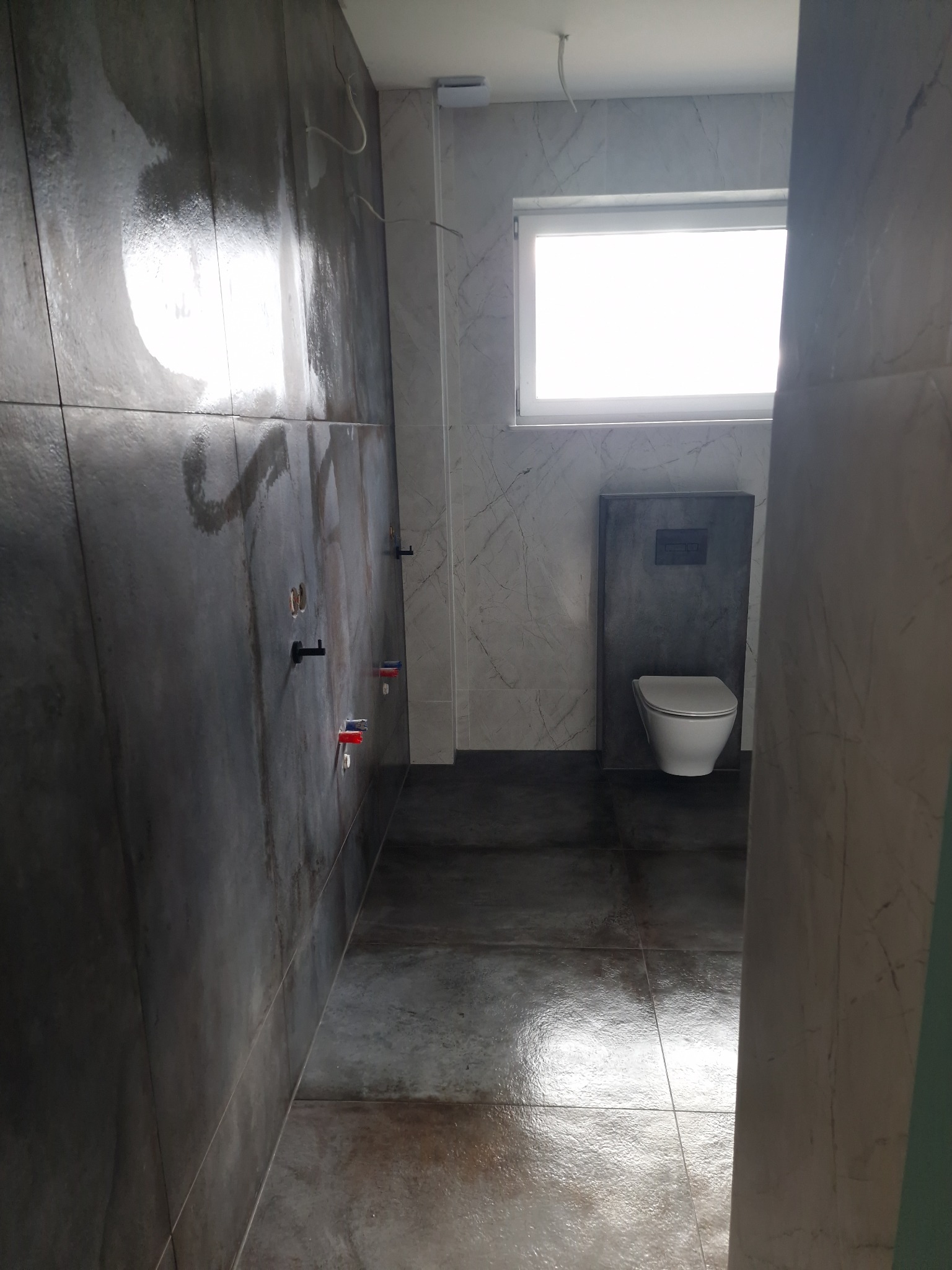 Wnętrze łazienki w trakcie prac wykończeniowych, widoczne płytki imitujące beton na ścianach z wyprowadzonymi instalacjami wodnymi oraz częściowo ułożona podłoga z ciemnych płytek, w tle...