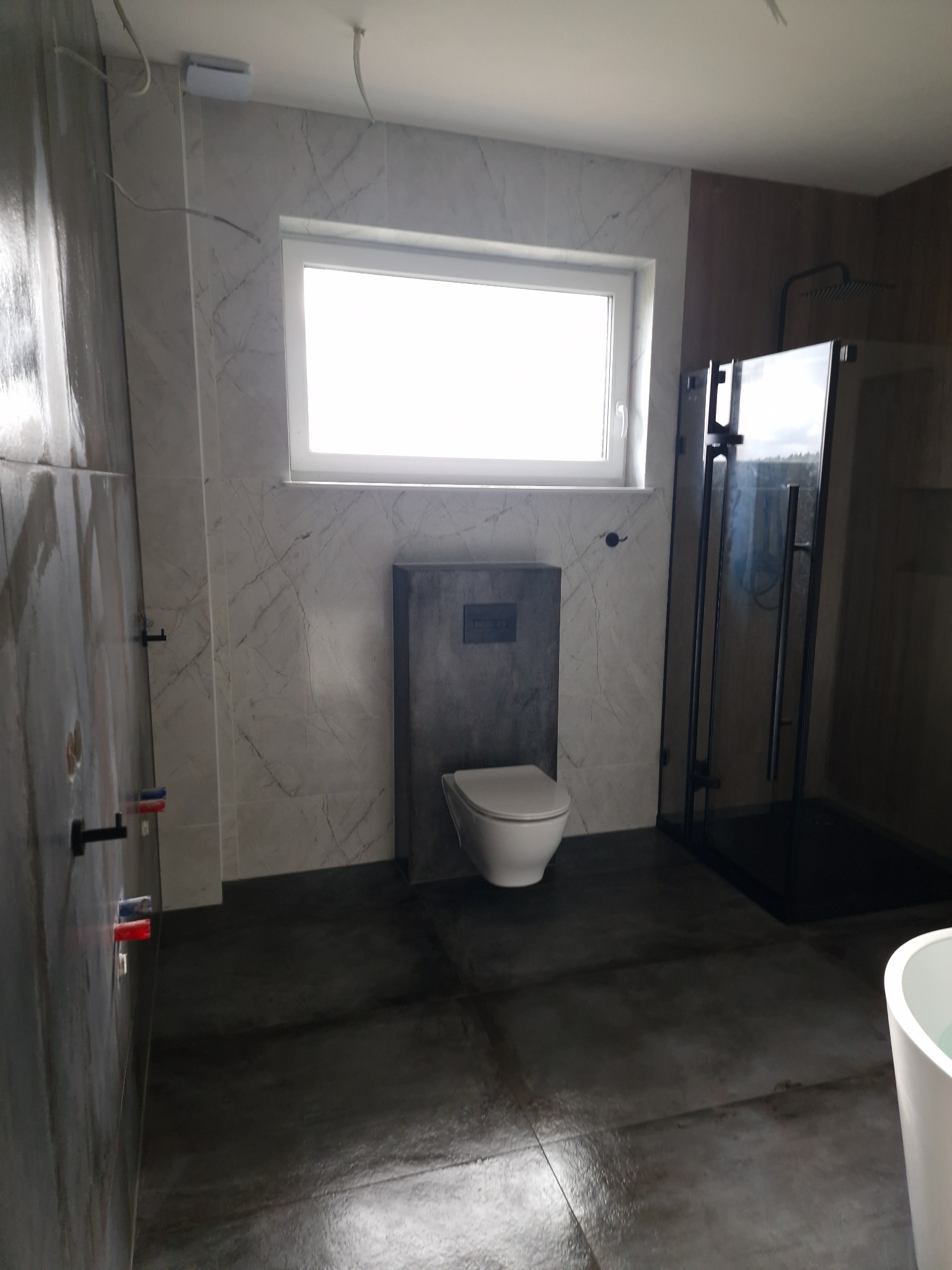 Nowoczesna łazienka po remoncie: wisząca toaleta z szarym panelem, kabina prysznicowa z czarnymi ramami, marmurowe płytki na ścianach, szara posadzka, widoczne przyłącza wodne na ścianie.