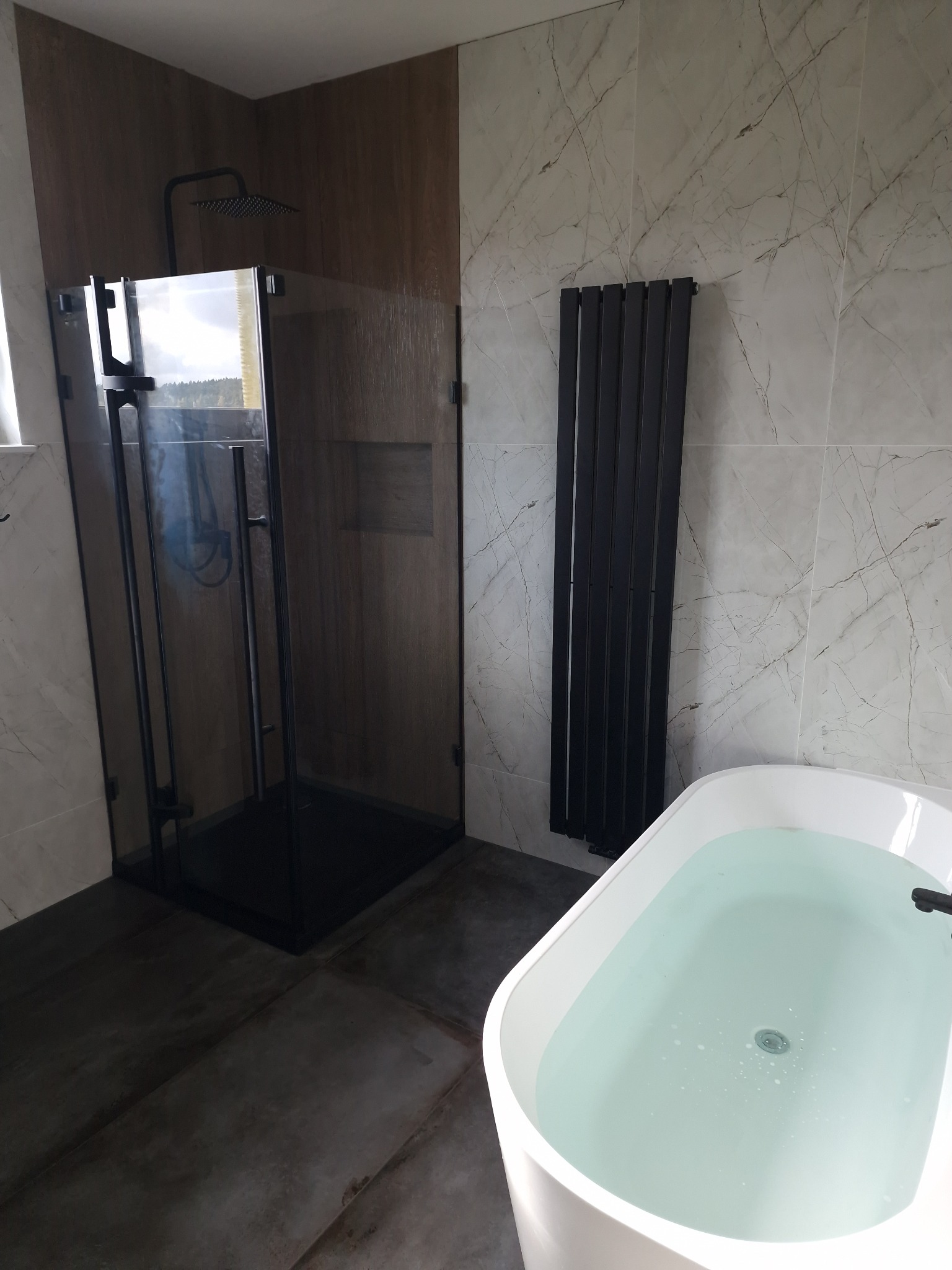 Nowoczesna łazienka z czarną kabiną prysznicową typu walk-in, grzejnikiem drabinkowym i białą wanną wypełnioną wodą, wykończona płytkami imitującymi marmur i betonową podłogą.