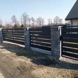 Nowoczesne ogrodzenie posesji z przesuwną bramą w kolorze grafitowym, z betonowymi słupkami, widok na posesję i dom w tle.