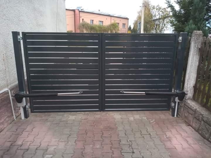Nowoczesna, pozioma brama dwuskrzydłowa z automatyką, szara, z widocznymi siłownikami, na tle kostki brukowej i zabudowań.