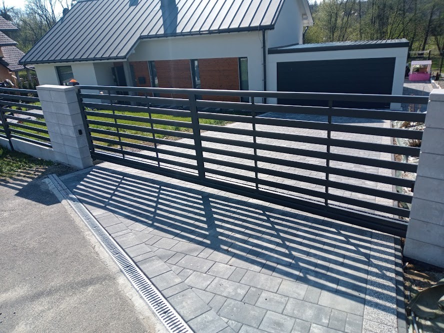 Nowoczesna, przesuwna brama wjazdowa z poziomymi, metalowymi profilami, osadzona na betonowych słupkach, z widocznym systemem odprowadzania wody liniowego i brukowaną kostką podjazdu, na tle domu...