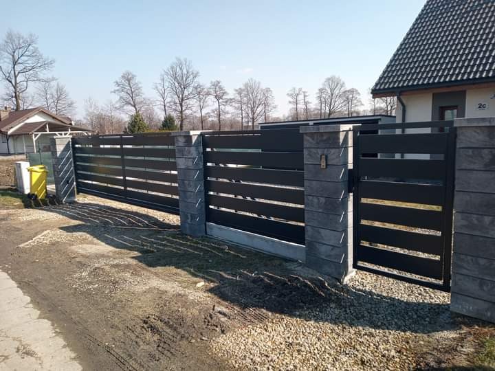 Nowoczesne ogrodzenie posesji z przesuwną bramą w kolorze grafitowym, z betonowymi słupkami, widok na posesję i dom w tle.