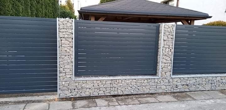 Nowoczesne ogrodzenie z poziomych, szarych paneli metalowych, z filarami i podstawą z gabionów wypełnionych jasnymi kamieniami, na tle zieleni i fragmentu budynku.