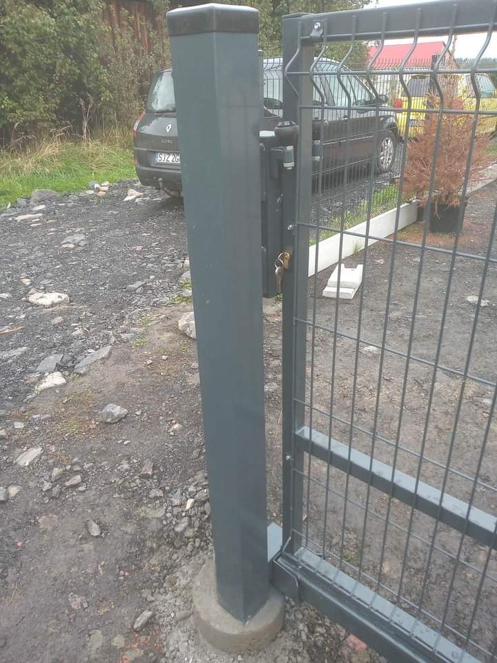 Fragment nowo zamontowanej, grafitowej bramy z siatki, z zamkiem i kluczami w zamku, na tle żwirowego podłoża i zaparkowanego samochodu osobowego.
