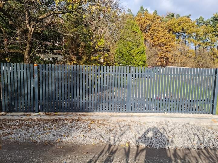 Nowoczesne, grafitowe ogrodzenie metalowe z pionowych profili, widok z zewnątrz, z bramą przesuwną i widocznymi drzewami w tle.