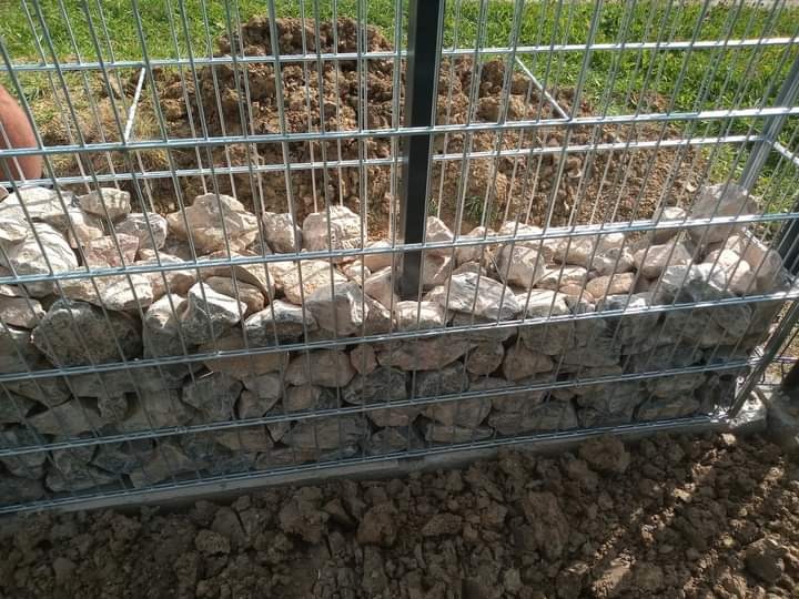 Fragment ogrodzenia gabionowego w trakcie budowy, wypełnionego jasnymi kamieniami, widoczne metalowe kosze z siatki i ziemia w tle.