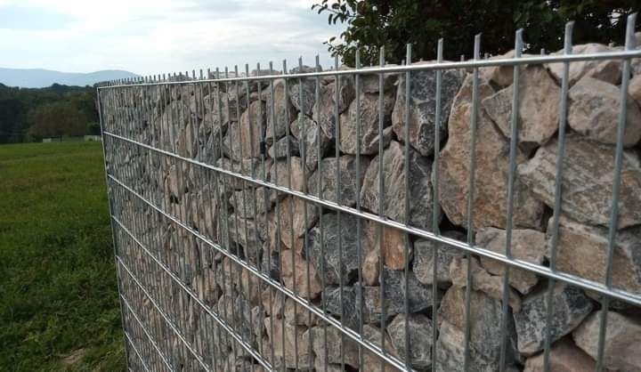 Gabionowa konstrukcja ogrodzeniowa z kamieniami, widok na zielone wzgórza i drzewa w oddali, metalowa siatka z wypełnieniem kamiennym.