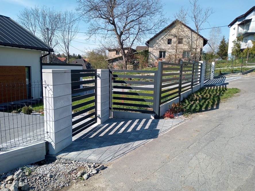 Nowoczesne ogrodzenie z metalowymi przęsłami i betonowymi słupkami w jasny dzień, widok z zewnątrz posesji, na pierwszym planie cień rzucany przez ogrodzenie na chodnik.