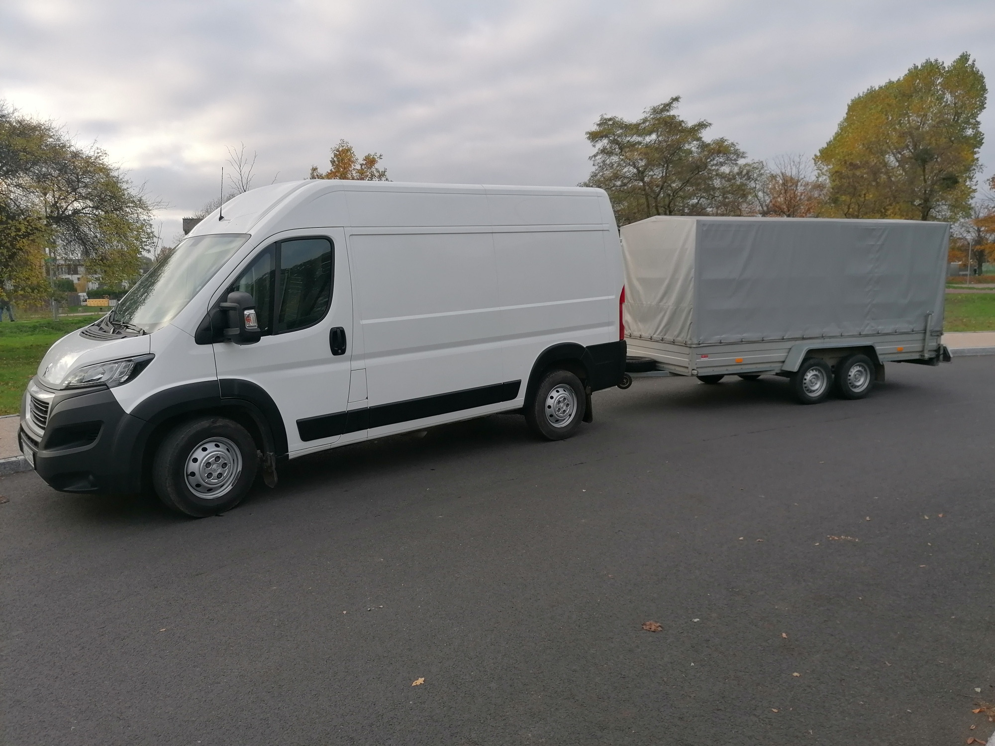 Biały samochód dostawczy z przyczepą krytą szarą plandeką na asfaltowej drodze, w tle drzewa z jesiennymi liśćmi pod pochmurnym niebem.