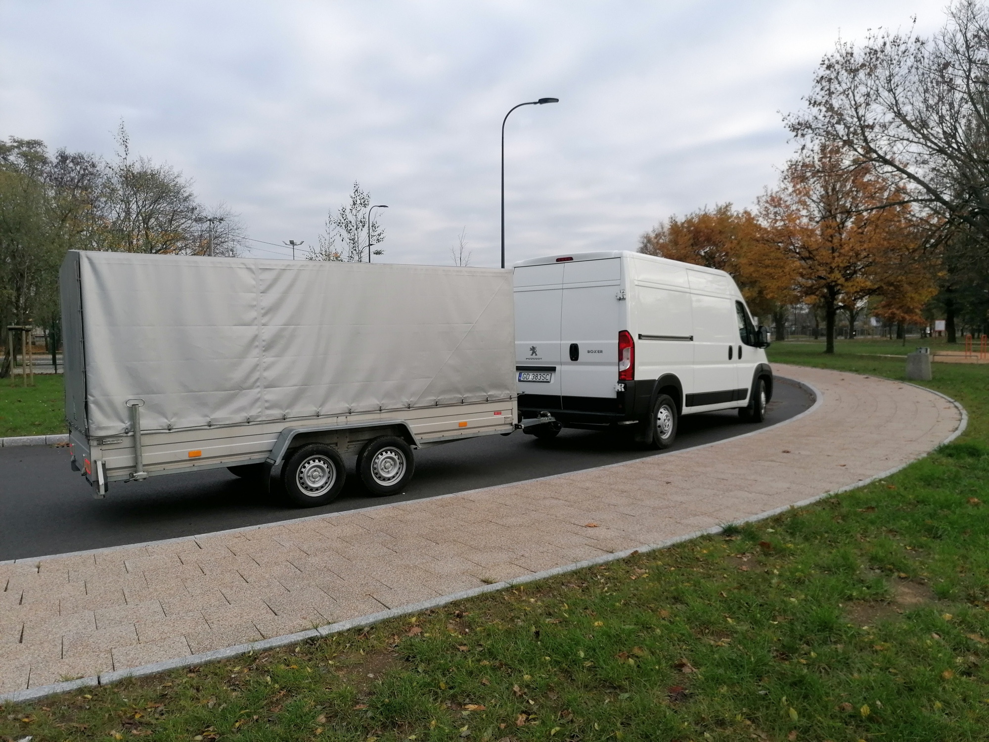 Biały samochód dostawczy Peugeot Boxer z przyczepą z plandeką, zaparkowany na ścieżce w parku, widok z tyłu.