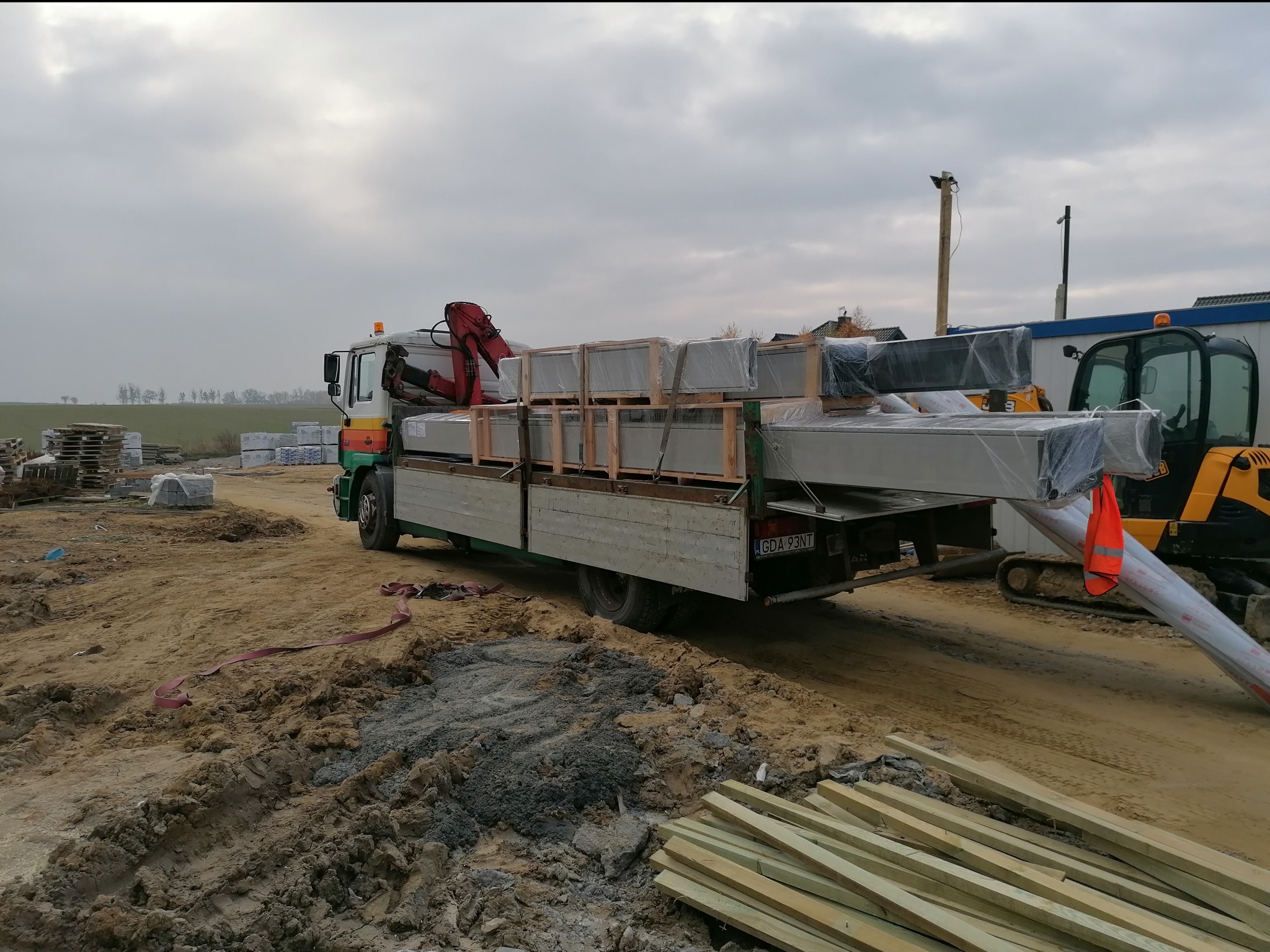 Ciężarówka z czerwoną ręką dźwigu załadowana paletami z zapakowanymi elementami, stojąca na placu budowy obok żółtej mini-koparki, z widocznymi stertami drewna i kostki brukowej w tle.