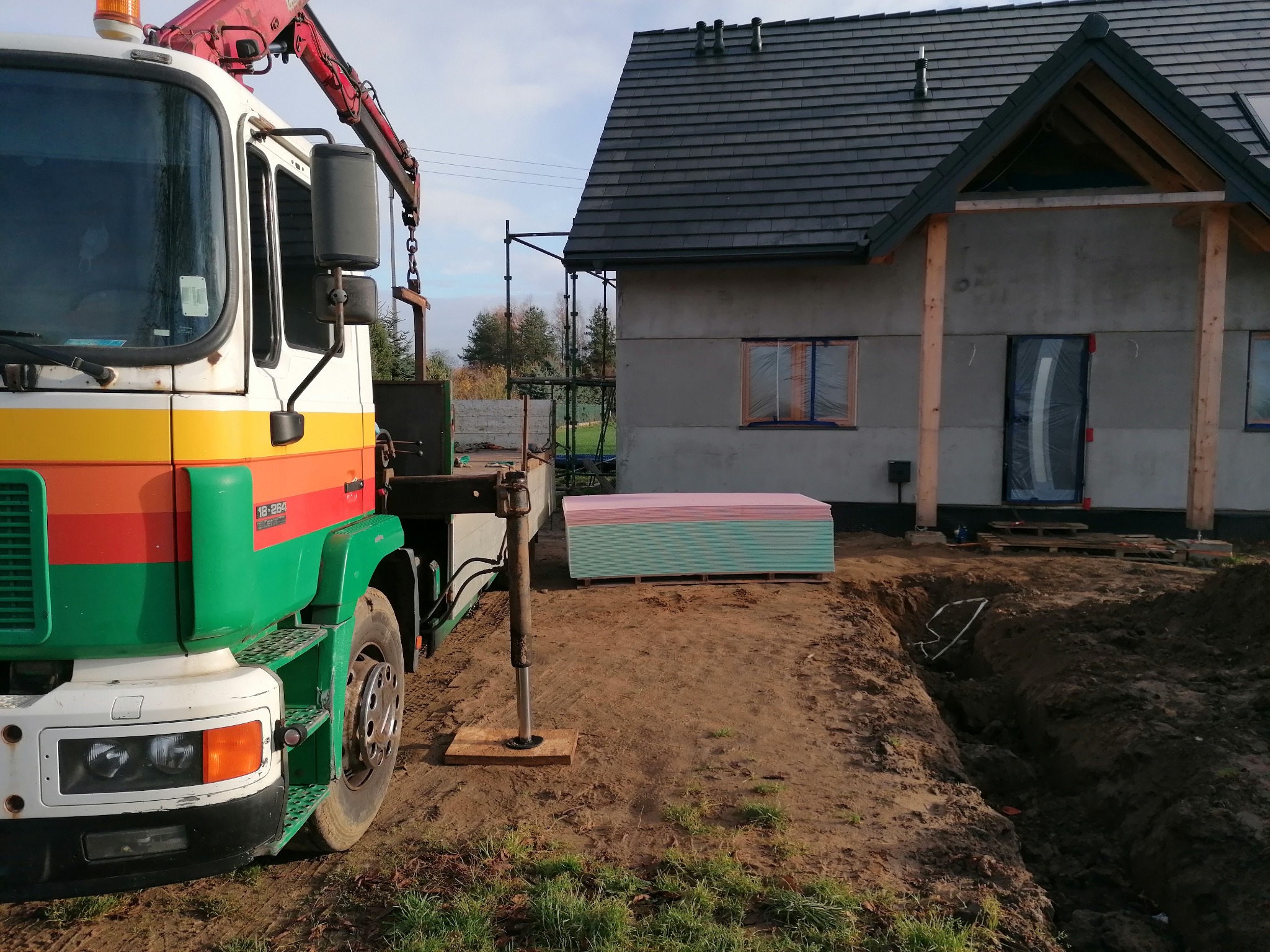 Żuraw samochodowy z podpartą hydraulicznie stopą stabilizującą, obok stos płyt gipsowo-kartonowych na palecie, w tle budynek w stanie surowym zamkniętym z czarną dachówką