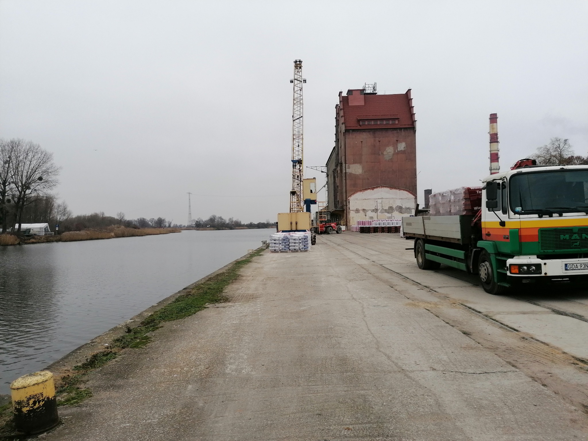 Ciężarówka MAN z zielono-pomarańczowym pasem załadowana paletami z materiałami budowlanymi stoi na betonowym nabrzeżu w porcie, w tle dźwig portowy i ceglany budynek magazynowy pod pochmurnym niebem.