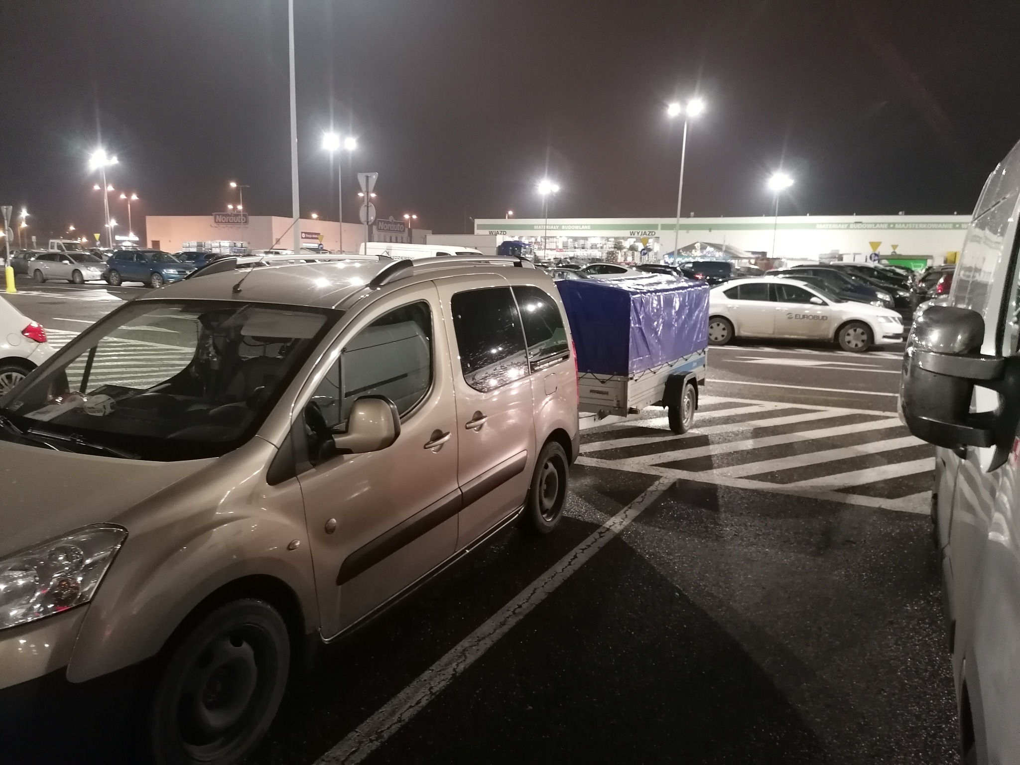 Beżowy samochód dostawczy z przyczepką pod plandeką na parkingu sklepu budowlanego o zmierzchu, widoczne oświetlenie uliczne.