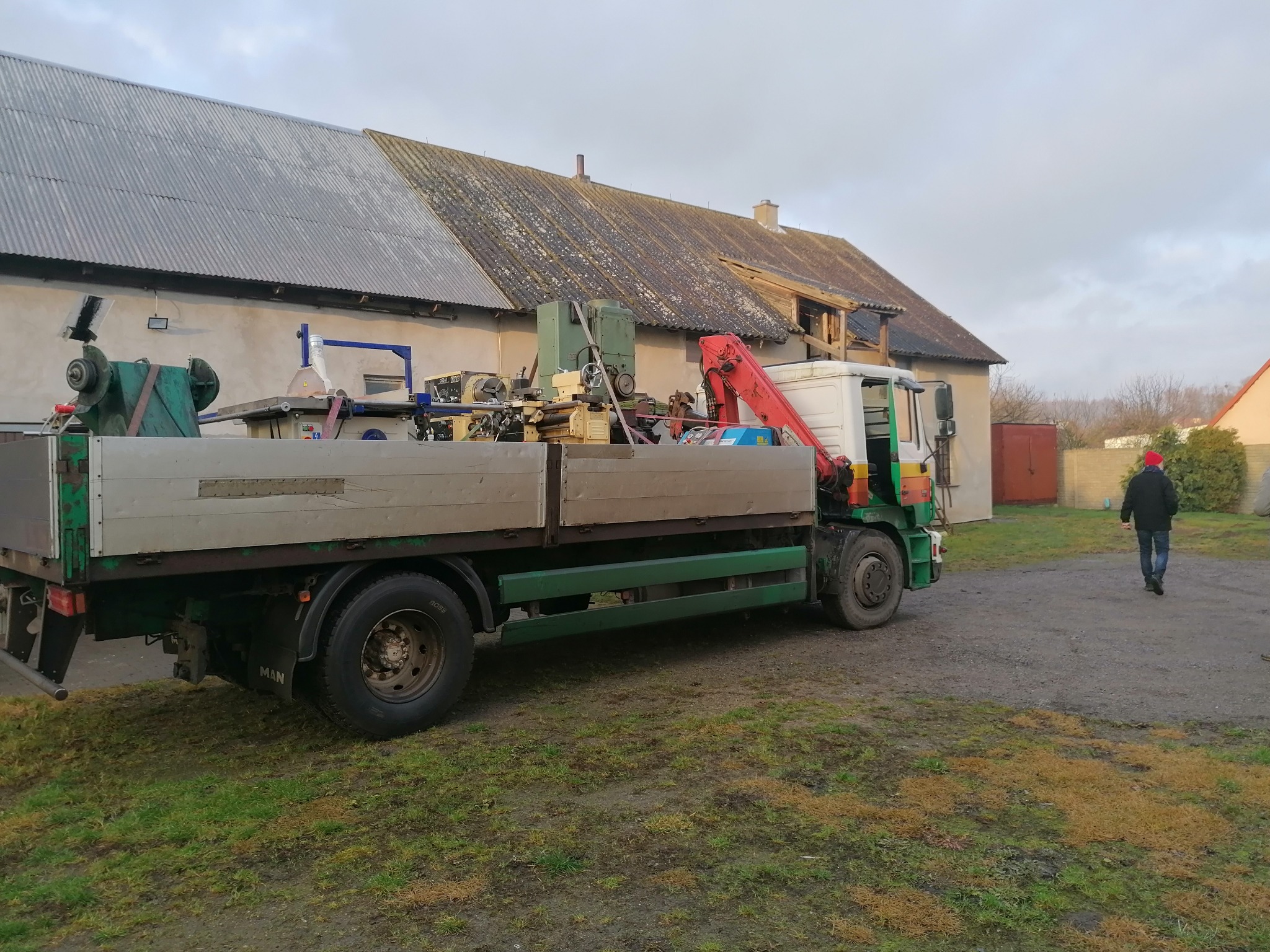 Ciężarówka MAN z podnośnikiem i różnymi maszynami na platformie, zaparkowana w Pruszczu Gdańskim na tle budynku gospodarczego i osoby oddalającej się.