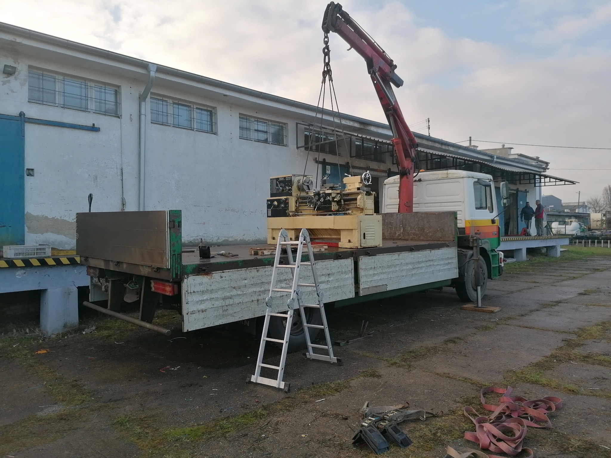 Ciężarówka z platformą załadowczą i dźwigiem transportuje tokarkę przemysłową; drabina oparta o platformę, budynek magazynowy w tle.