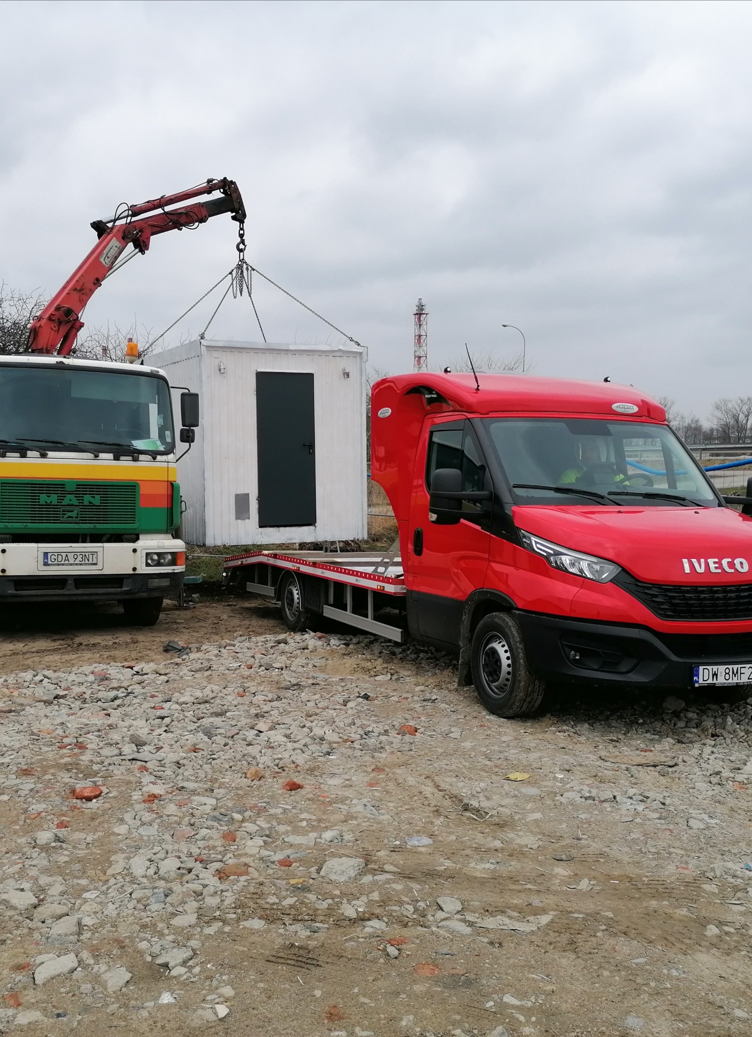 Ciężarówka MAN z kolorowym pasem na grillu i czerwona ciężarówka Iveco na gruzowisku, dźwigiem podnoszona biała budka z czarnymi drzwiami.