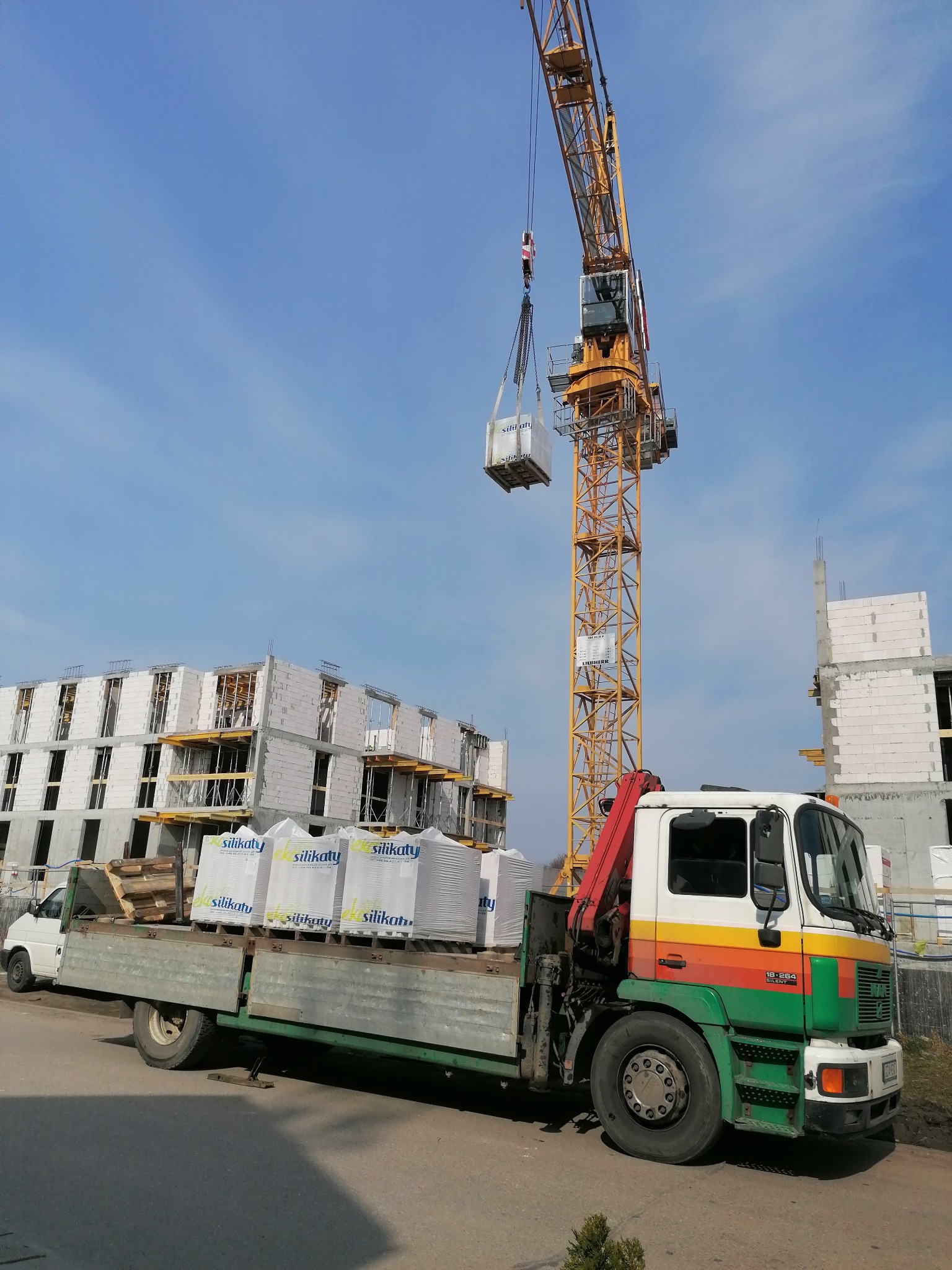 Żółty dźwig podnosi pakiet białych bloczków silikatowych nad zielono-pomarańczowo-białą ciężarówką na tle budynku w trakcie budowy z bloczków silikatowych i błękitnego nieba.