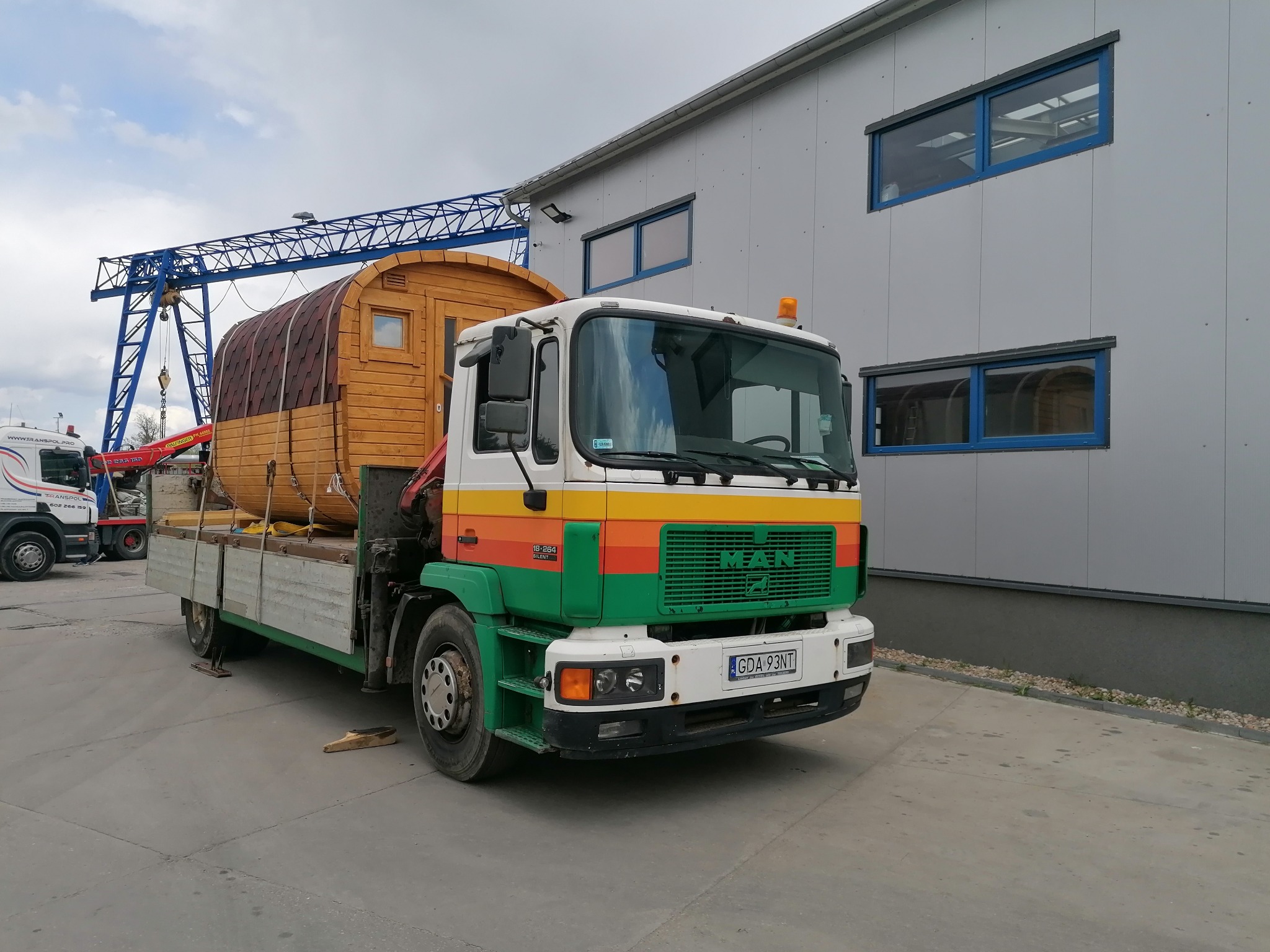 Ciężarówka MAN z zielono-pomarańczowym pasem transportuje drewnianą saunę beczkę na platformie, w tle widoczny suwnicowy żuraw i szara hala przemysłowa.